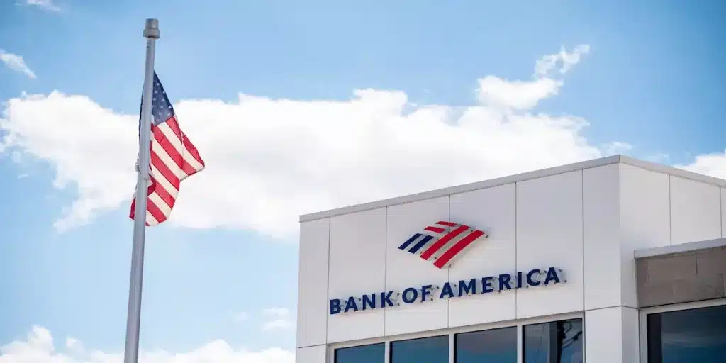 Exterior of Bank of America building showing red blue white logo and BANK OF AMERICA text, with American flag waving on pole under partly cloudy sky