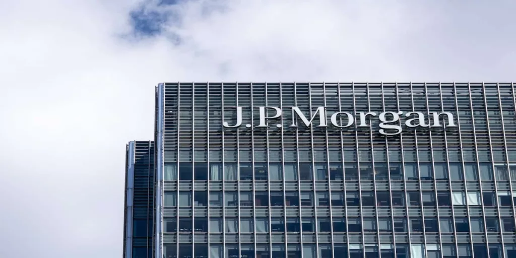 Close-up of J.P. Morgan white logo mounted on facade of modern glass and steel office tower against blue sky with clouds